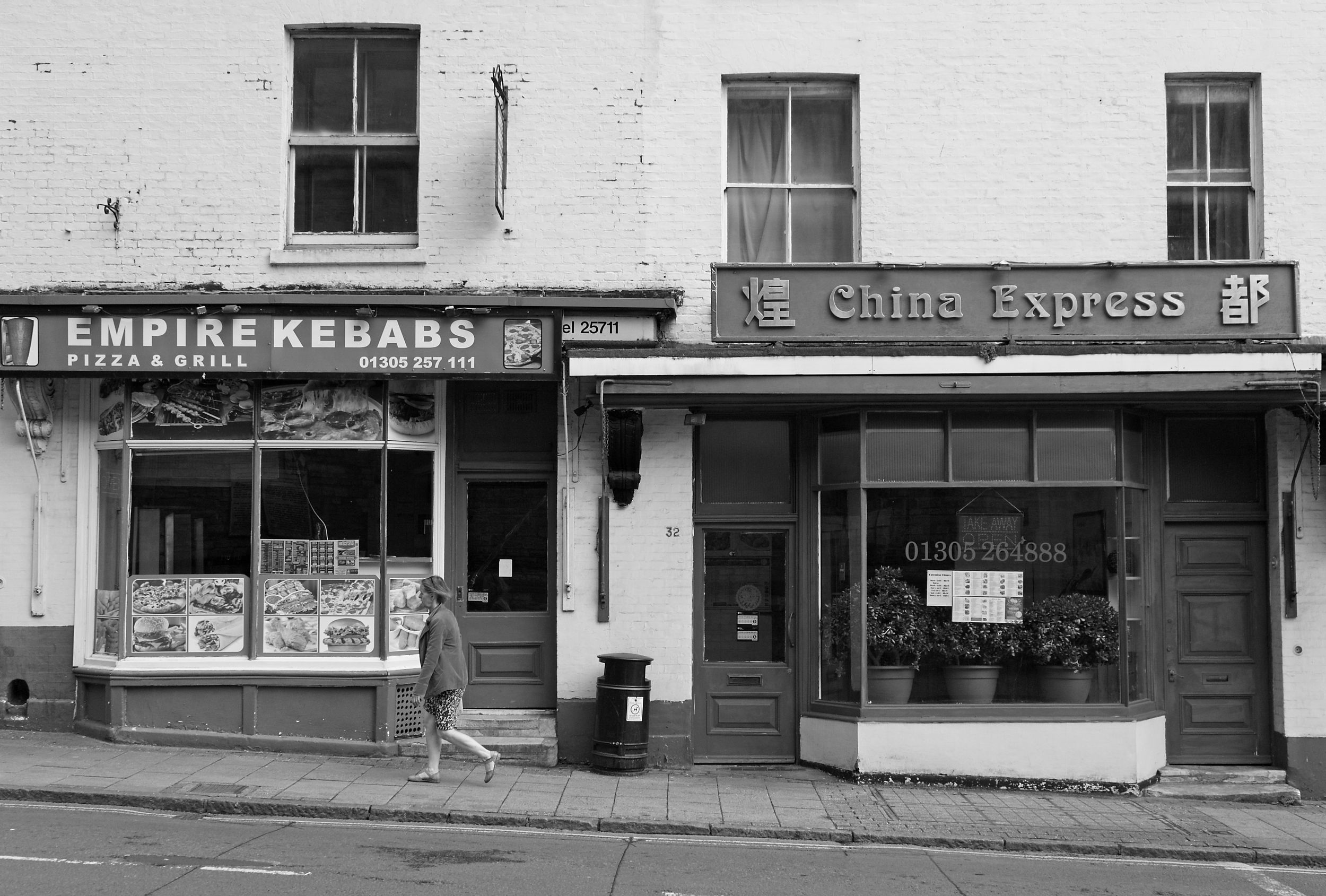 Black and white image of 2 take away food shops as a woman walks in fromt of them.